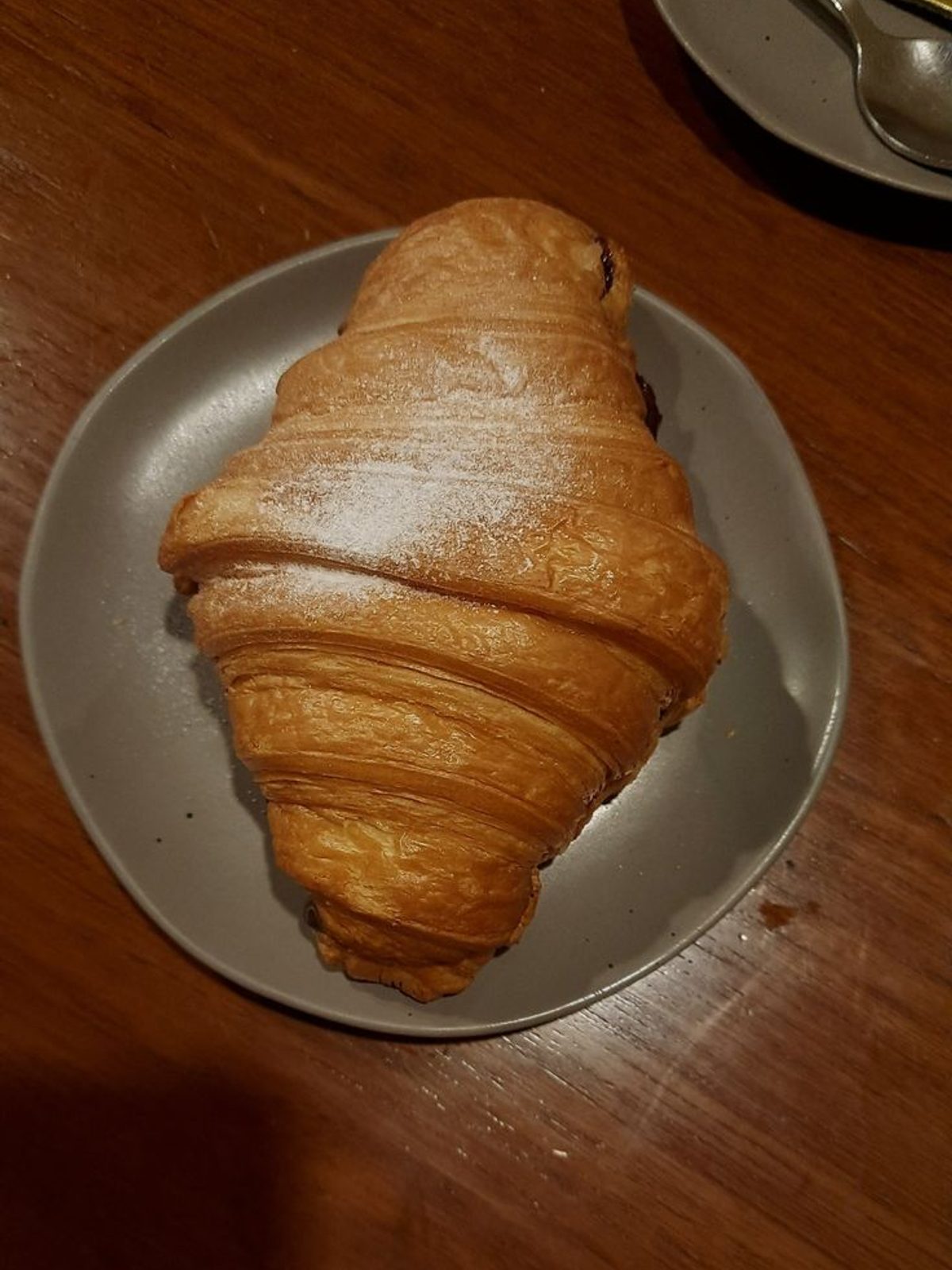 Phone photo of a croissant under flat cafe light, looking like a smooth brown crescent with no visible flake